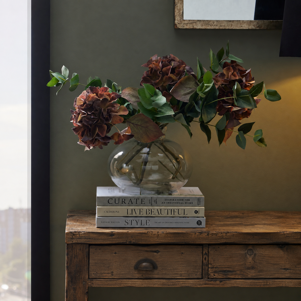 A clear glass vase filled with dried burgundy hydrangea flowers and green eucalyptus leaves, displayed on a stack of books on a rustic wooden console table against a dark olive wall.
