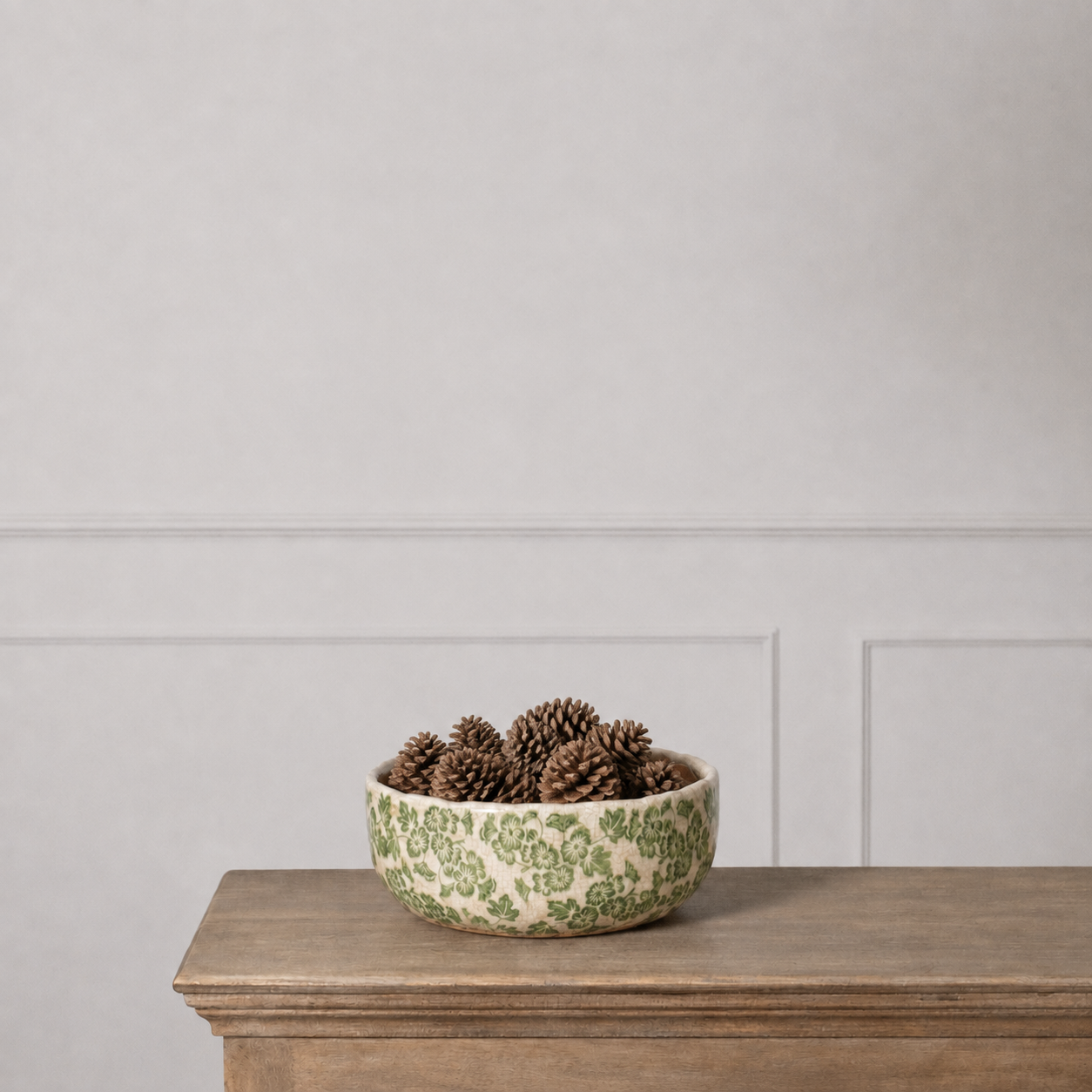 A ceramic bowl with a green floral pattern on a cream background, filled with pinecones and placed on a wooden mantelpiece against a light gray paneled wall.