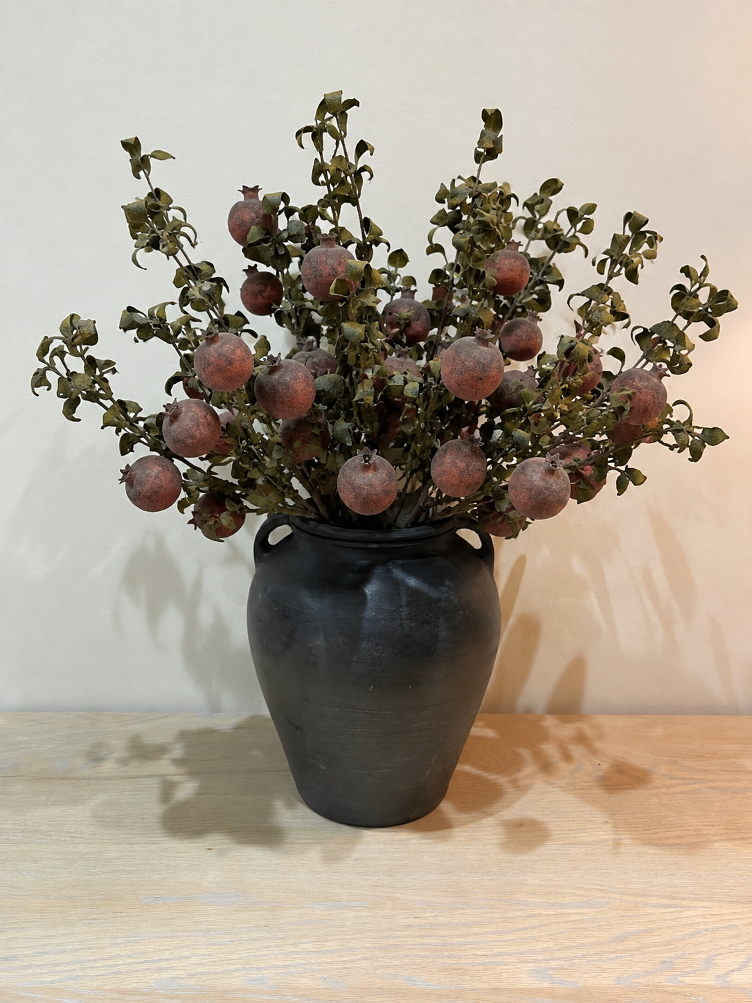 A decorative artificial plant arrangement with small round dusty-rose fruits and green-brown leaves, displayed in a dark matte ceramic vase on a light wood surface against a plain off-white wall.