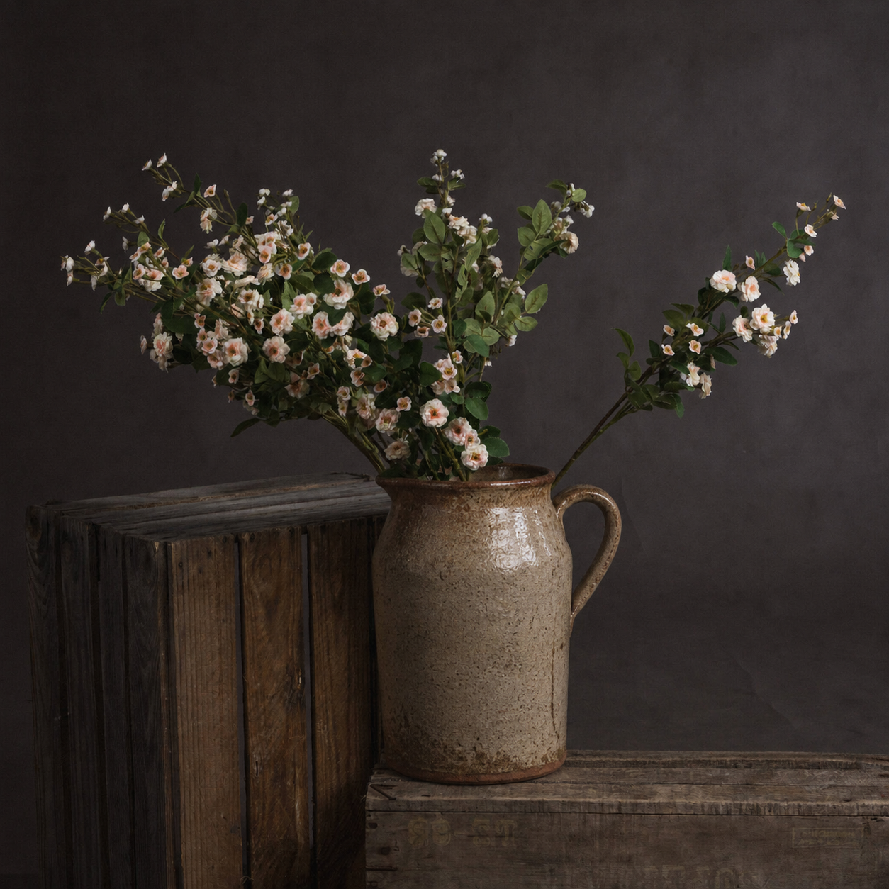 A rustic ceramic pitcher vase filled with white and pink wildflowers, set on a wooden surface against a dark gray background.