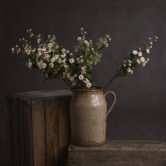 A rustic ceramic pitcher vase filled with white and pink wildflowers, set on a wooden surface against a dark gray background.