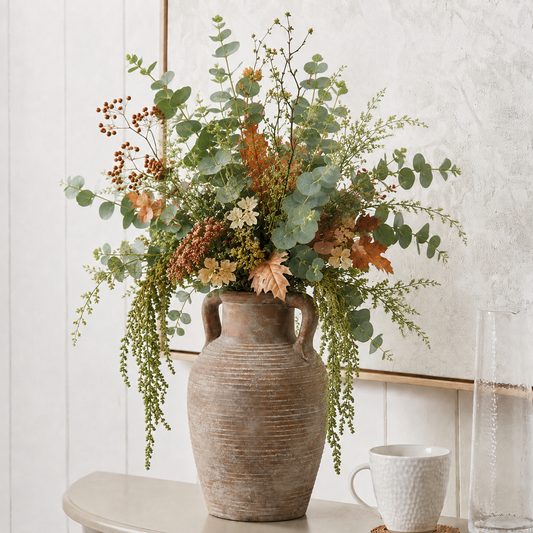 A rustic brown ceramic vase filled with a lush arrangement of green eucalyptus leaves and warm autumnal florals, displayed on a light-colored table against a textured white wall.