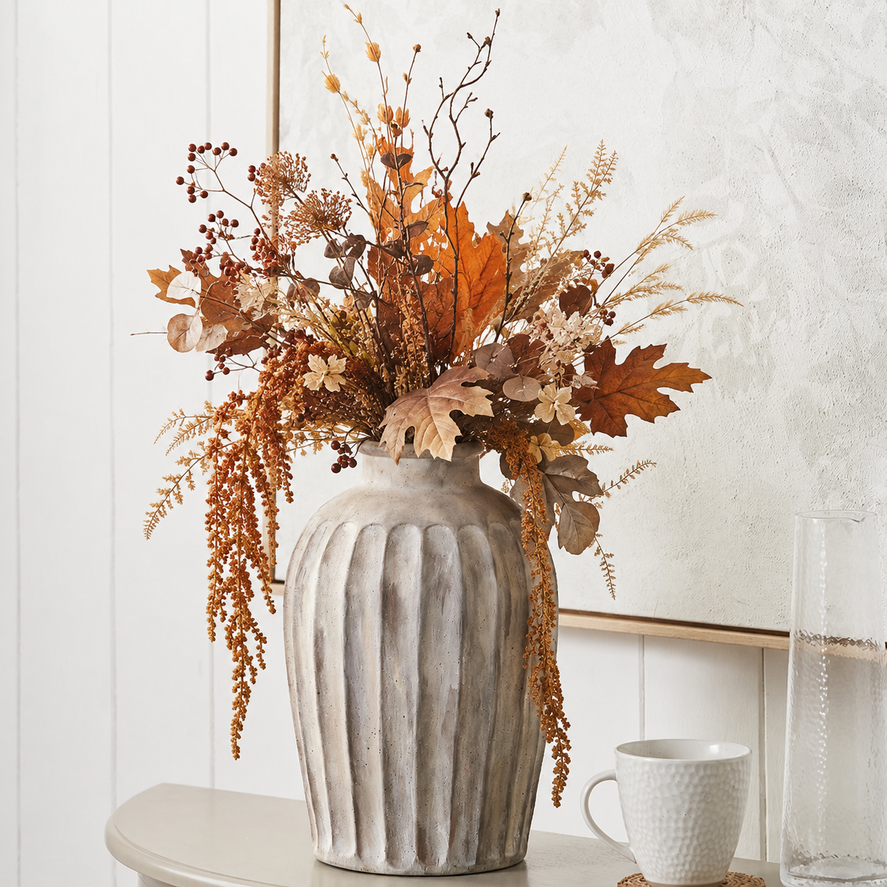 An autumn floral arrangement in a ribbed gray ceramic vase, displayed on a tabletop against a white paneled wall with a white textured mirror and a white textured mug nearby.
