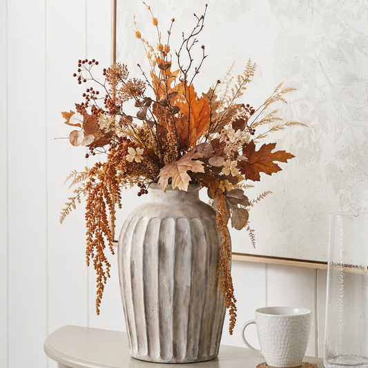 An autumn floral arrangement in a ribbed gray ceramic vase, displayed on a tabletop against a white paneled wall with a white textured mirror and a white textured mug nearby.